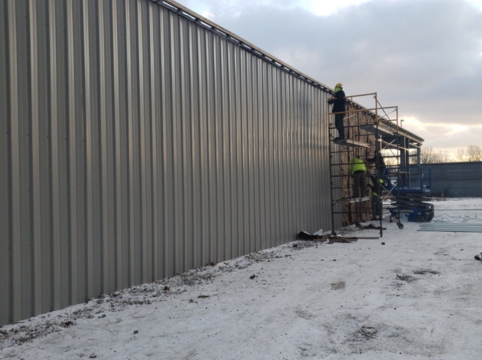 Two workers in yellow vests fix an exterior wall of a gray corrugated metal building on scaffolding. scaffolding, construc...